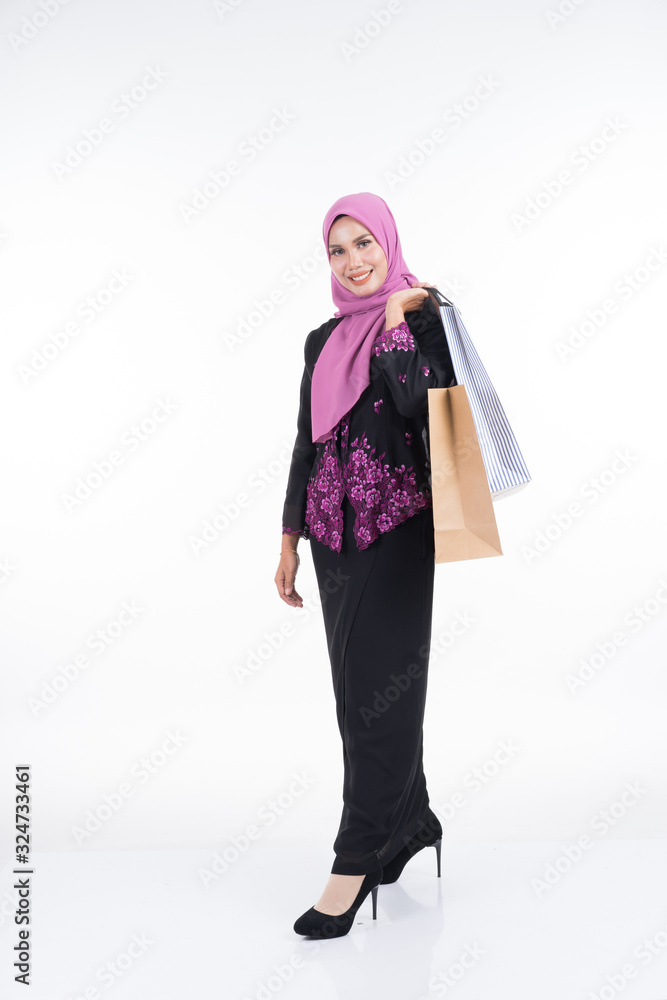 A beautiful Asian female model in a traditional dress kebaya carrying shopping bags isolated on white background. Eidul fitri festive preparation shopping concept. Full length portrait. A beautiful Asian female model in a traditional dress kebaya carrying shopping bags isolated on white background. Eidul fitri festive preparation shopping concept. Full length portrait.