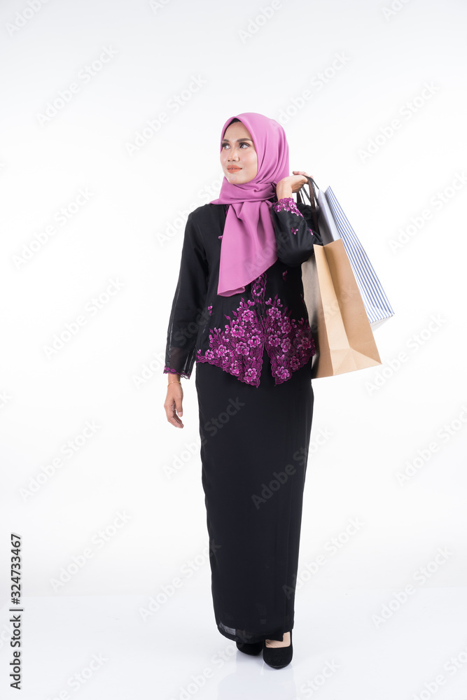 A beautiful Asian female model in a traditional dress kebaya carrying shopping bags isolated on white background. Eidul fitri festive preparation shopping concept. Full length portrait.