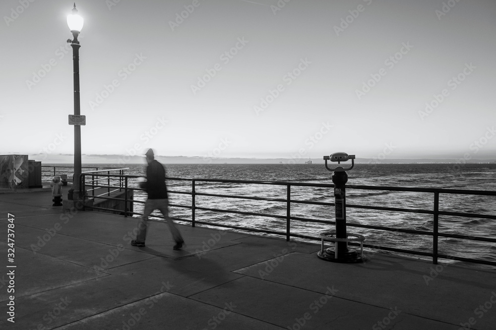 Landscape of the Pacific Ocean as a man walks briskly on the Huntington Beach pier during twilight.