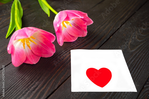 Fototapeta Naklejka Na Ścianę i Meble -  Two fresh pink tulip flowers and white sheet of paper with heart on top lie on dark wooden background. Happy holiday, womens day or wedding greetings concept. Copy space for text.