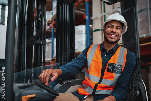 Fotografie, Obraz Young forklift driver sitting in vehicle in warehouse smiling looking at camera