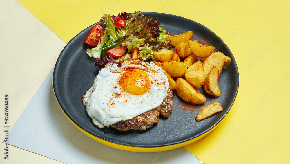 Beef steak with fried eggs with a side dish of salad and fried potatoes on a black plate on a colored background. Appetizing food for lunch