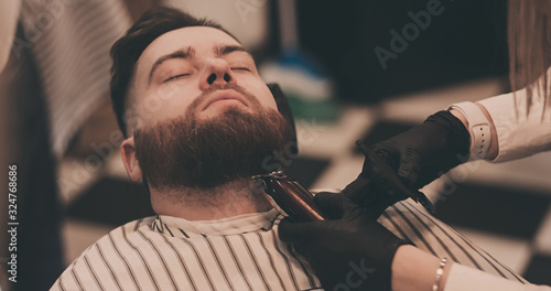Barber cutting beard of male client. Hairstylist serving client at barber shop.