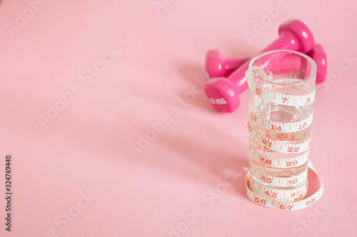 pink dumbbells, glass of fresh water and measuring tape in inches on pink background, weight loss concept