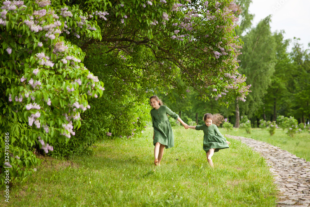 Fototapeta premium Two sisters in green linen dress have fun in the park with blooming lilacs, enjoy spring and warmth