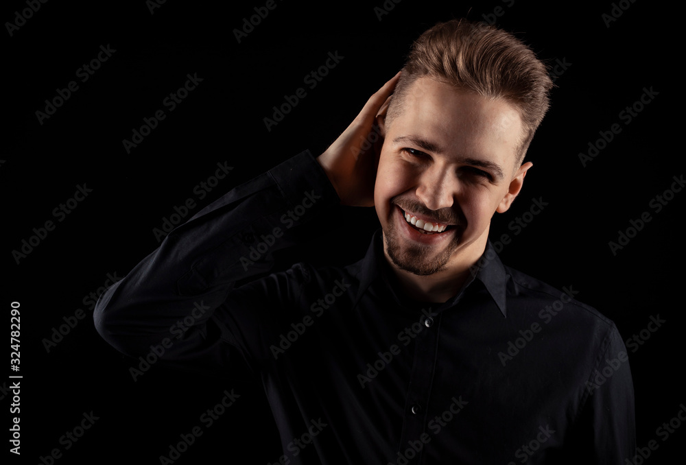 Cheery young man with goatee in black shirt touching hair with one hand looking at camera, isolated