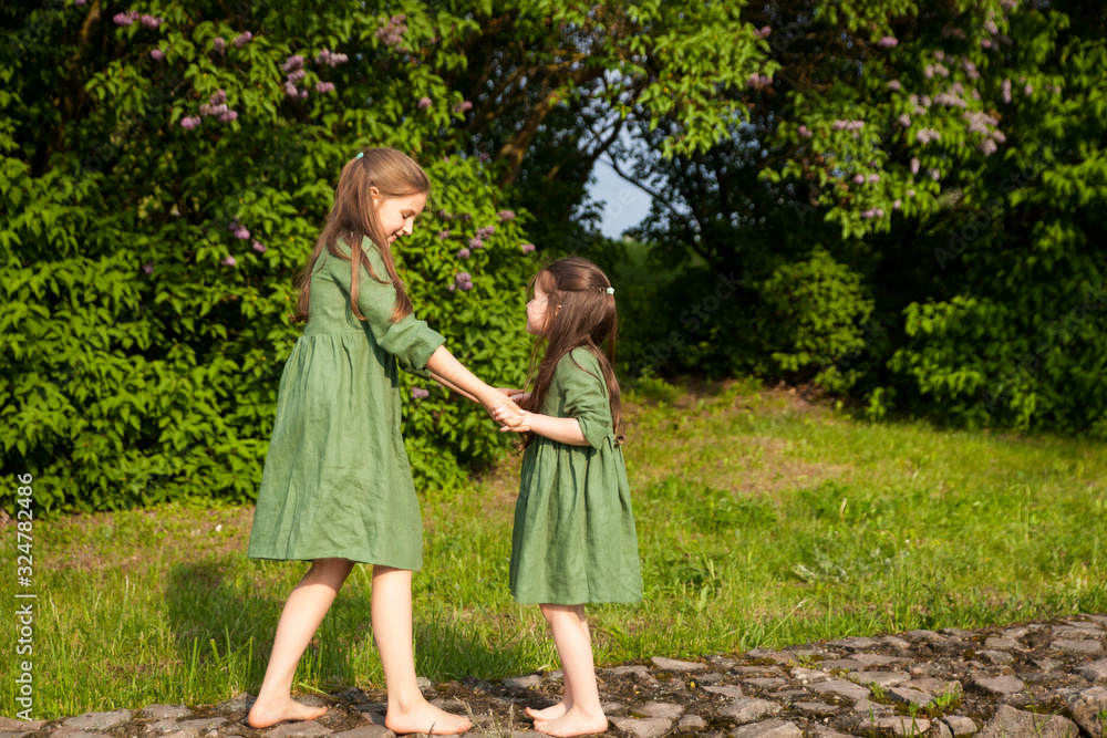 Two sisters in green linen dress have fun in the park with blooming lilacs, enjoy spring and warmth Two sisters in green linen dress have fun in the park with blooming lilacs, enjoy spring and warmth