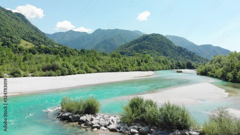 Drone flies over pebble beach and turquoise clear mountain river with rolling green hills in background, sunny summer day in Slovenia, Europe. Drone flies over pebble beach and turquoise clear mountain river with rolling green hills in background, sunny summer day in Slovenia, Europe.