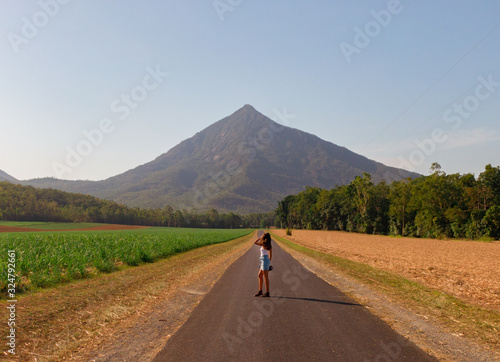  Woman & Beautiful mountain landscape. Sugar cane fields foreground. Dramatic DRONE aerial view of fields, trees, green forest, farm, mountains & road. Shot in Walsh's Pyramid, Cairns, Australia.