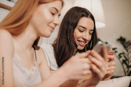 Charming caucasian girl with red hair and freckles is texting with her brunette friend sitting on a sofa