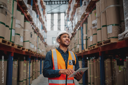 Low angle view of young african man wearing reflective jacket holding digital tablet standing in factory warehouse smiling photo