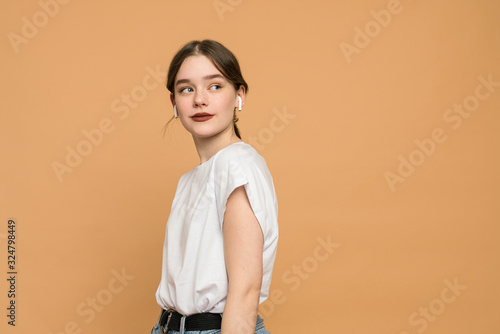 Closeup of a young brunette woman in wireless earphones dressed in white t-shirt, student listening to the music and looking away isolated over orange background with copyspace