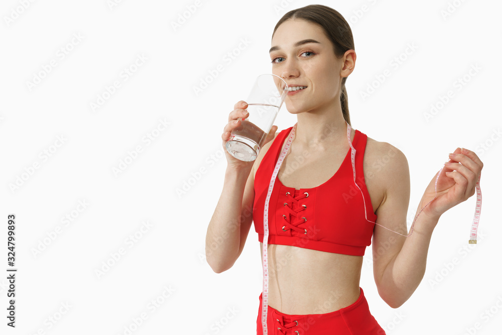 Attractive female in red sportswear drink water, looking at the camera and holds tape measure in her hand, isolated over white background Attractive female in red sportswear drink water, looking at the camera and holds tape measure in her hand, isolated over white background