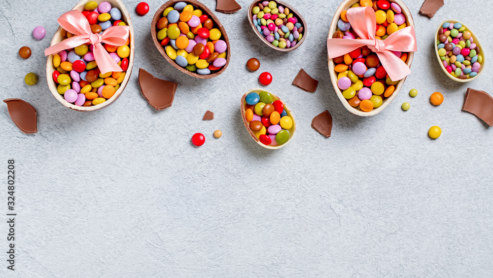 Easter border with chocolate eggs and colorful candy sweets on grey concrete table, copy space. Traditional Easter treats flat lay, holiday background, web banner