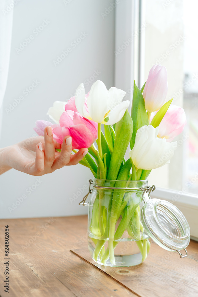 Woman hand touching buds of tulips in a vase on windowsill