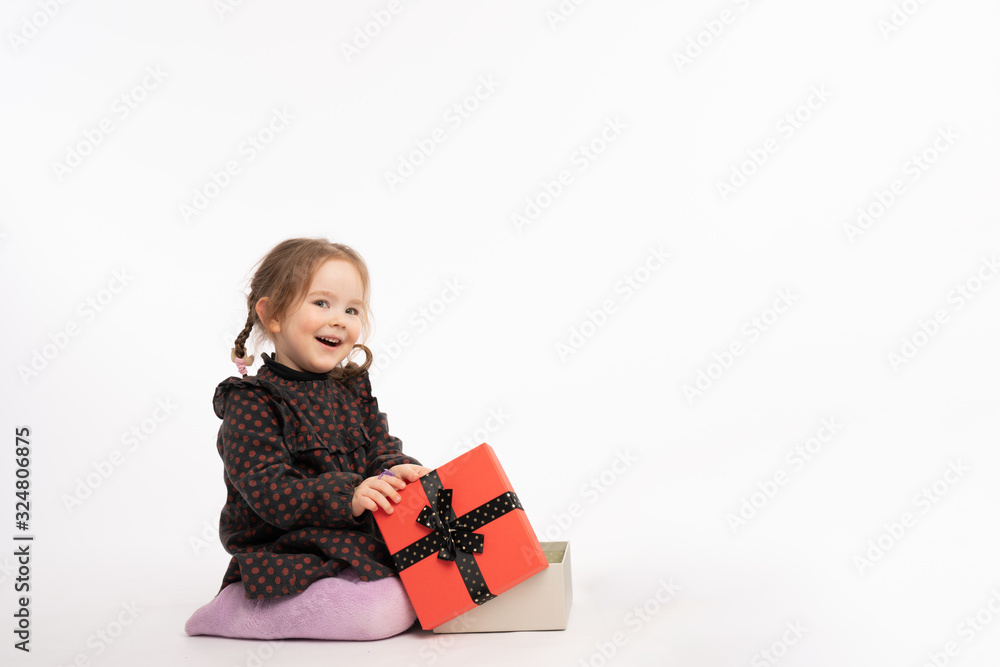 Happy smiling redhead girl in purple tights and red pea dress sitting on the floor with New Year's gift and looking at the camera, isolated over white background with copyspace Happy smiling redhead girl in purple tights and red pea dress sitting on the floor with New Year's gift and looking at the camera, isolated over white background with copyspace