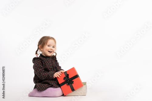 Happy smiling redhead girl in purple tights and red pea dress sitting on the floor with New Year's gift and looking at the camera, isolated over white background with copyspace