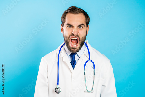 Angry doctor man in white coat screaming to camera. Stressed and depressed man on blue background.