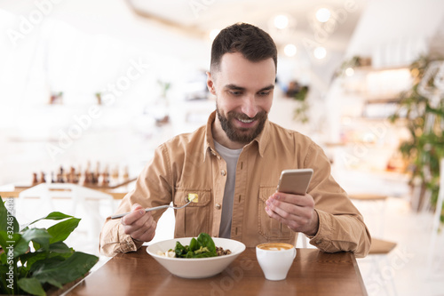 Young bearded smiling man drinks his hot coffee and eats salad for lunch while checking news in his smartphone during break   multitasking concept