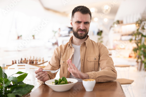 Young bearded man seems to be unhappy with his salad during lunch at the cafe  complaints and suggestions concept