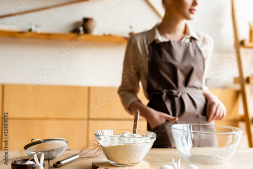 cropped view of woman in apron standing with hand in pocket near bowls and ingredients