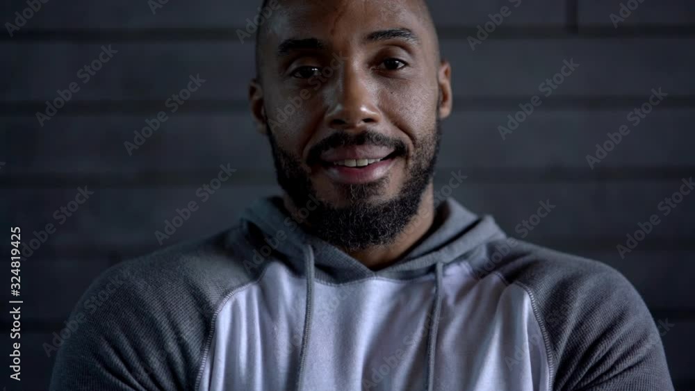 Muscular ethnic bald sportsman in sportswear with white earphones smiling and looking at camera while listening to music near grey stone wall Muscular ethnic bald sportsman in sportswear with white earphones smiling and looking at camera while listening to music near grey stone wall