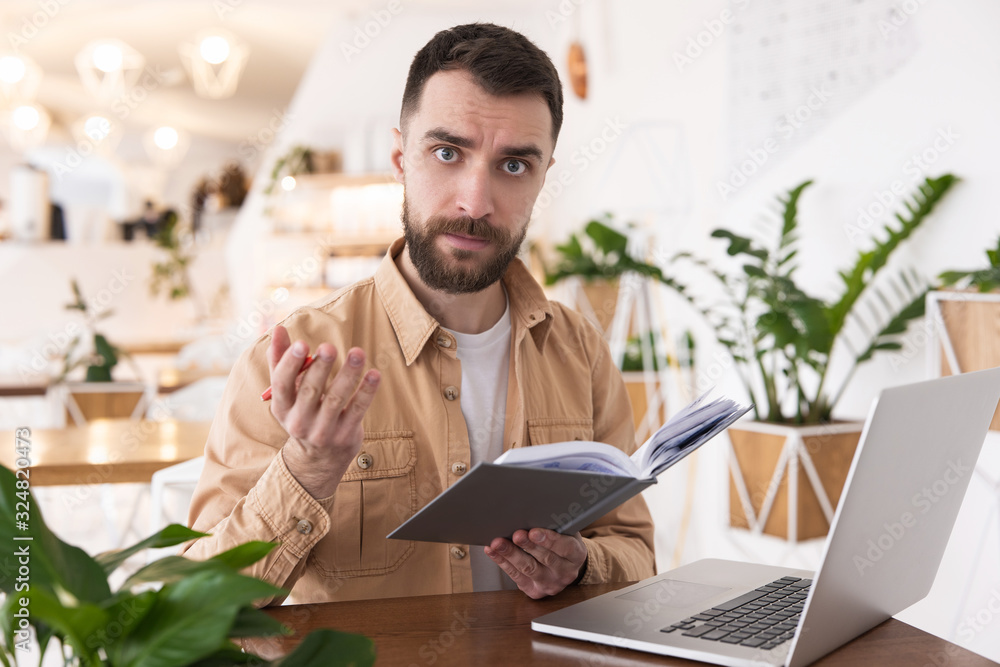 Young handsome bearded man looks resolute working in laptop holding pen and planner during lunch break at cafe, multitasking concept Young handsome bearded man looks resolute working in laptop holding pen and planner during lunch break at cafe, multitasking concept