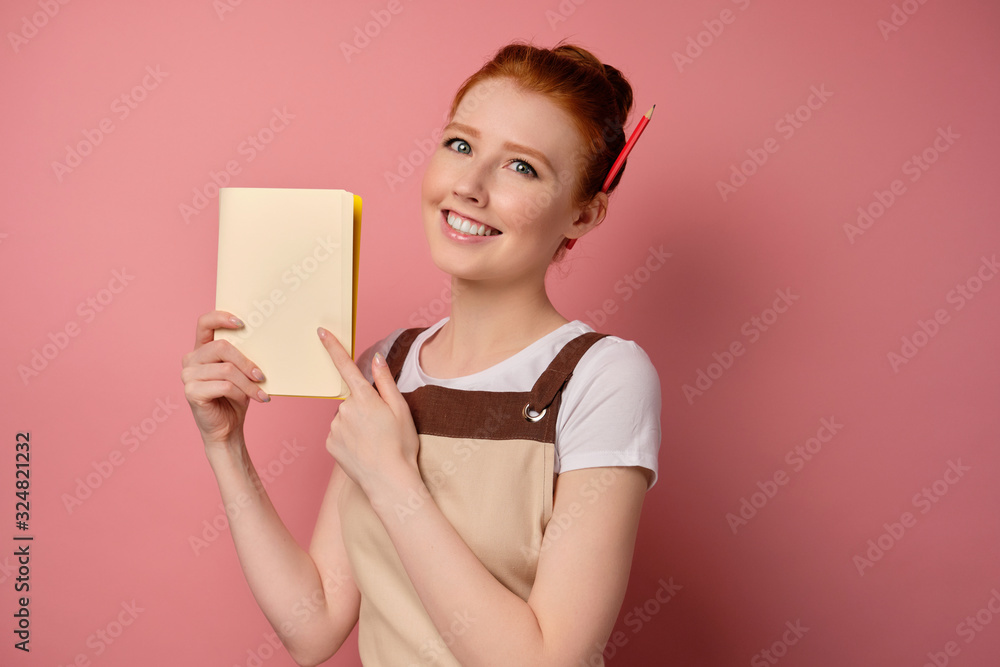 A cute red-haired girl with a high bun in a beige apron pointing to the notebook page with finger and smiling A cute red-haired girl with a high bun in a beige apron pointing to the notebook page with finger and smiling