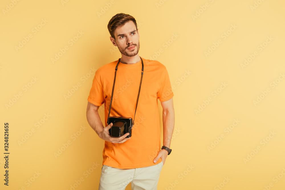 handsome thoughtful photographer with digital camera holding hand in pocket and looking away on yellow background handsome thoughtful photographer with digital camera holding hand in pocket and looking away on yellow background