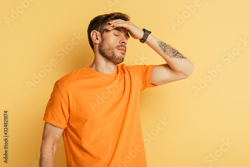 exhausted young man touching forehead while standing with closed eyes on yellow background