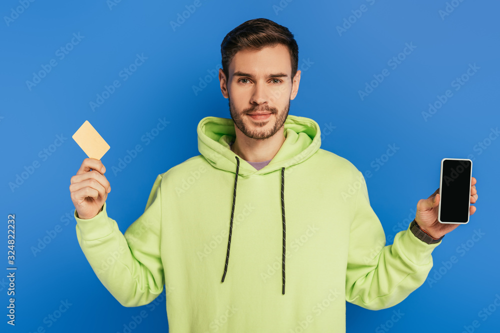 positive young man showing smartphone with blank screen and credit card isolated on blue positive young man showing smartphone with blank screen and credit card isolated on blue