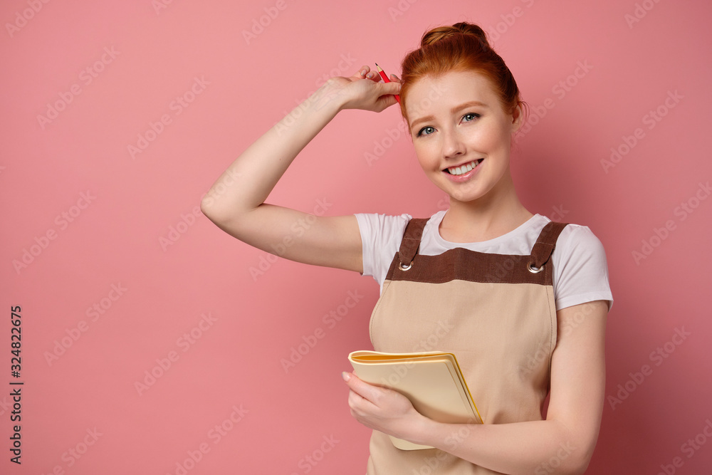 A cute red-haired girl with a high bun in a beige apron is smiling, holding a notebook to herself and putting a pencil behind ear A cute red-haired girl with a high bun in a beige apron is smiling, holding a notebook to herself and putting a pencil behind ear