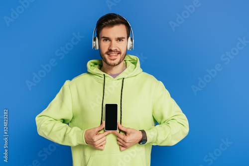 cheerful young man in wireless headphones showing smartphone with blank screen isolated on blue © LIGHTFIELD STUDIOS