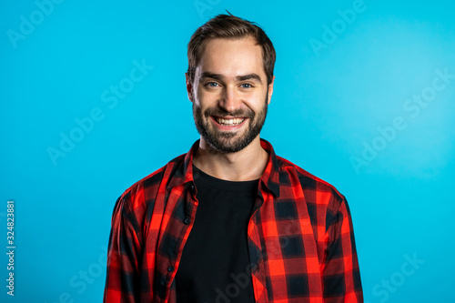Handsome european man with trendy beard in red plaid shirt on blue studio background. Cheerful guy smiling and looking to camera.
