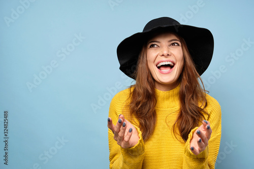 A charming brunette in a yellow sweater and hat stands on a blue background and laughs with his mouth wide open