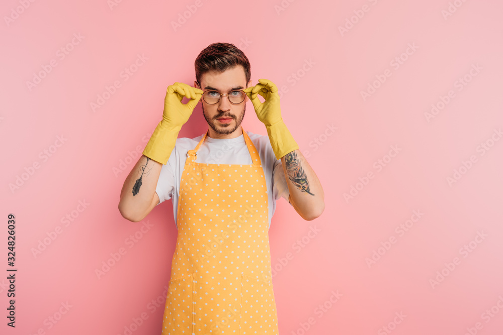 confident young man in apron and rubber gloves touching glasses on pink background confident young man in apron and rubber gloves touching glasses on pink background