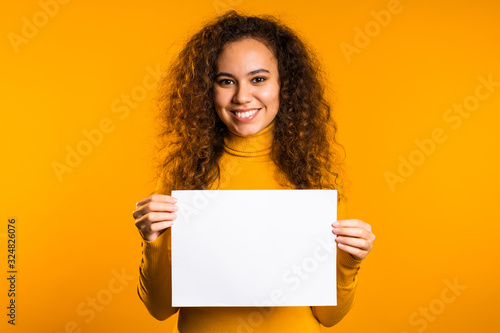Pretty curly woman holding horizontal white a4 paper poster. Copy space. Smiling trendy girl on yellow background. photo