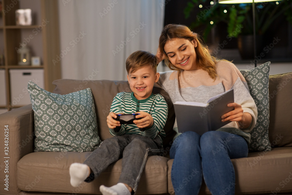 family, leisure and people concept - happy smiling mother reading book and her little son with gamepad playing video game in evening family, leisure and people concept - happy smiling mother reading book and her little son with gamepad playing video game in evening