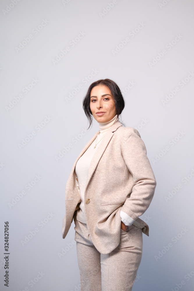 Portrait of a smiling mid-aged business woman in a light milk-colored trouser suit and turtleneck against a light wall. Portrait of a smiling mid-aged business woman in a light milk-colored trouser suit and turtleneck against a light wall.