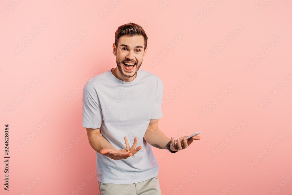 impressed young man showing wow gesture while holding smartphone on pink background impressed young man showing wow gesture while holding smartphone on pink background