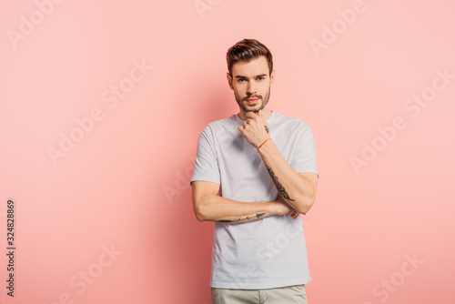 thoughtful young man touching chin while looking at camera on pink background