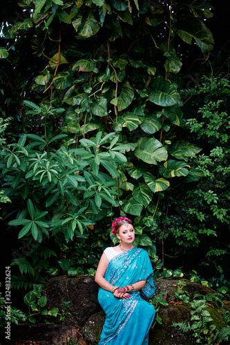 Beautiful young caucasian woman in traditional indian clothing with bridal makeup and jewelry. Bollywood dancer in Sari and henna on hands walking on green garden.