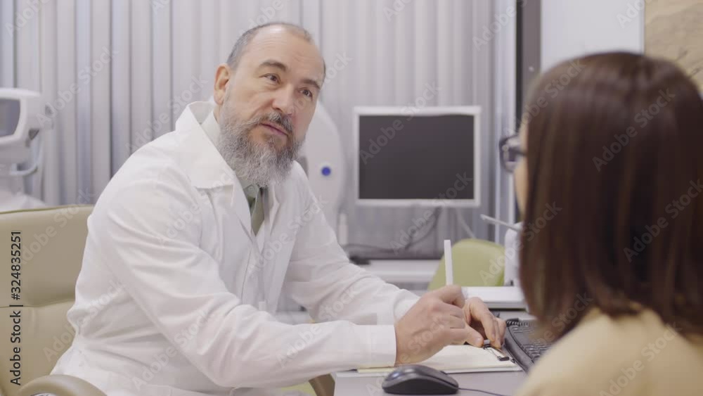 Senior doctor in lab coat sitting at desk and speaking with female patient during medical consultation in ophthalmology clinic Senior doctor in lab coat sitting at desk and speaking with female patient during medical consultation in ophthalmology clinic