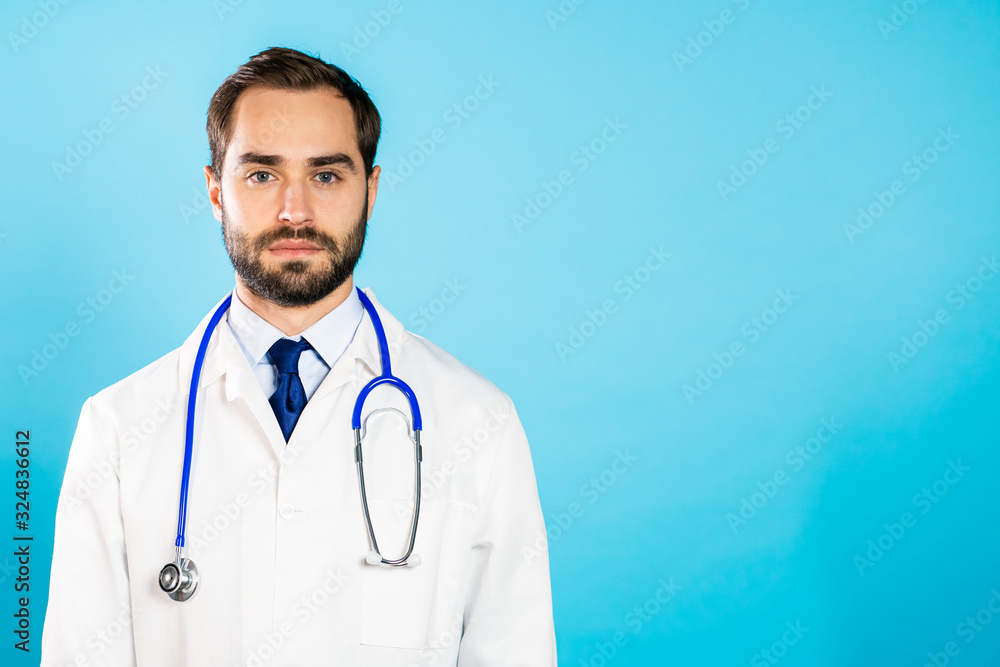 Portrait of young handsome man in professional medical white coat is isolated on blue studio background. Doctor with beard and stethoscope. Copy space Portrait of young handsome man in professional medical white coat is isolated on blue studio background. Doctor with beard and stethoscope. Copy space