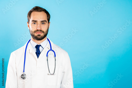 Portrait of young handsome man in professional medical white coat is isolated on blue studio background. Doctor with beard and stethoscope. Copy space