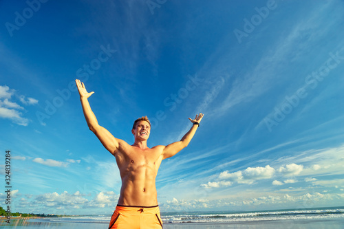 Victory and freedom. Young handsome strong man raising hands up on the beach against sky.