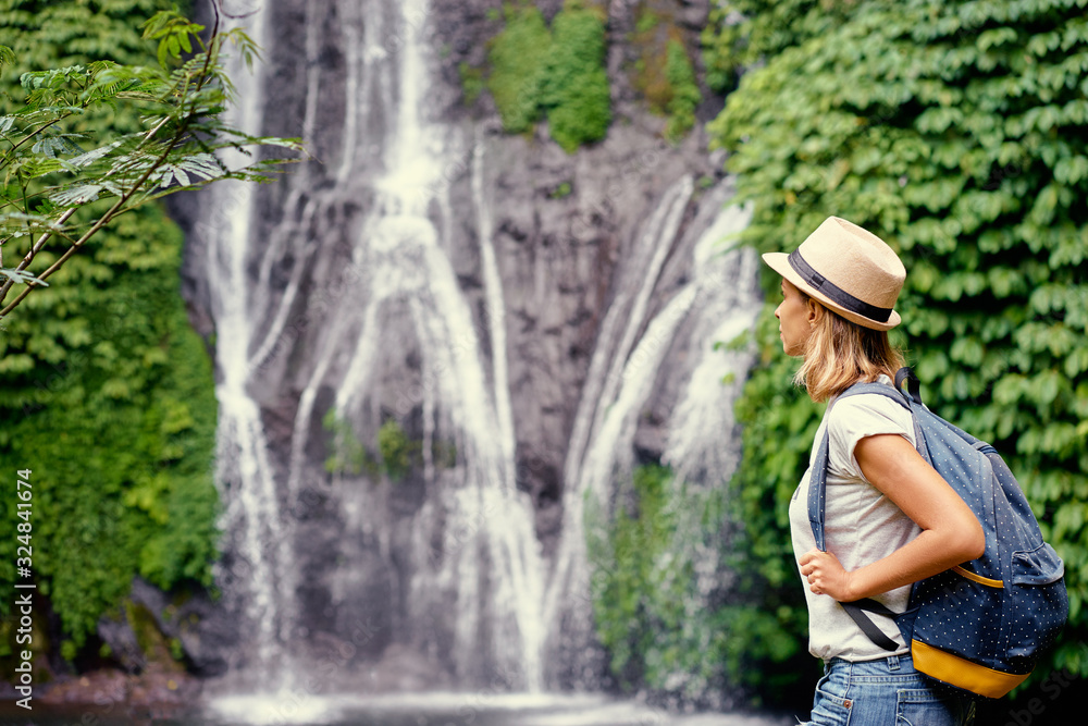 Tropical traveling. Young woman in hat with rucksack enjoying waterfall view.