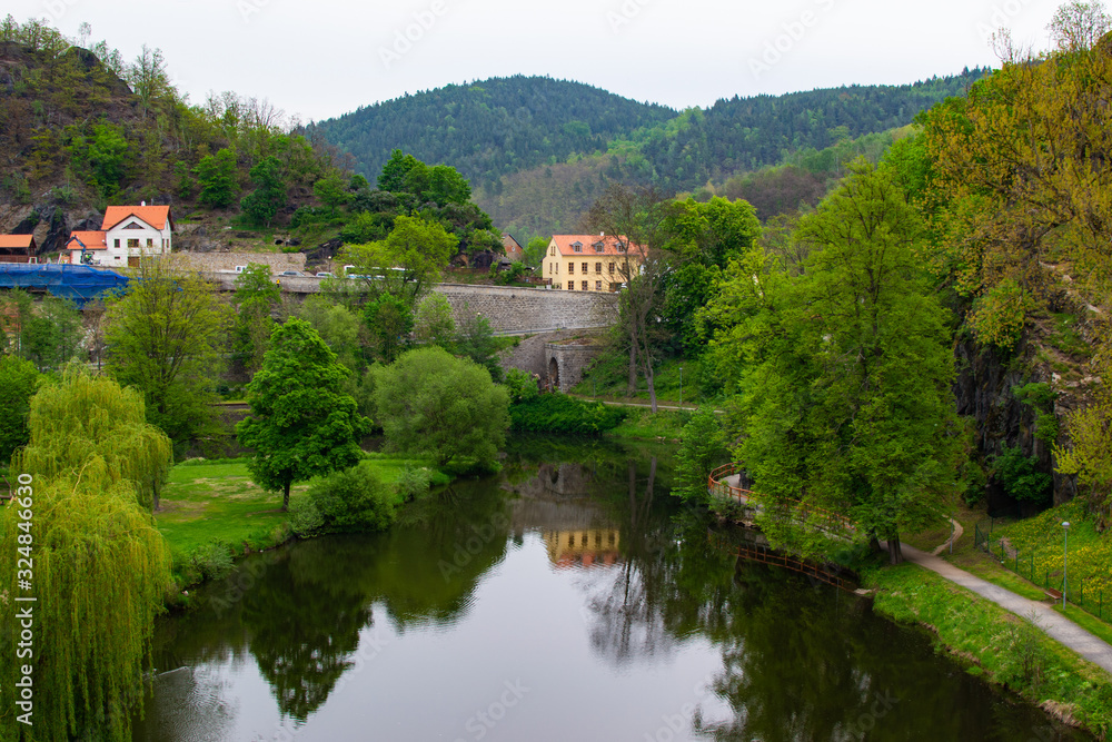 Reflections in the water of Ohre river between trees in Loket, Czech Republic, with typical houses, a stone wall and a bridge at the background