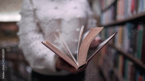  Unrecognizable woman beautifully turns over the pages of old paper book in big library on shelves background. Beautiful scene. Education, science, university concept.
