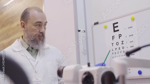 Senior optometrist in lab coat pointing at letters on eyechart with pencil while checking eyesight of patient photo